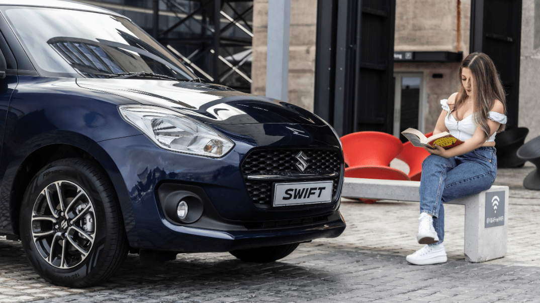 Black Suzuki Swift with lady reading book infront of the car.